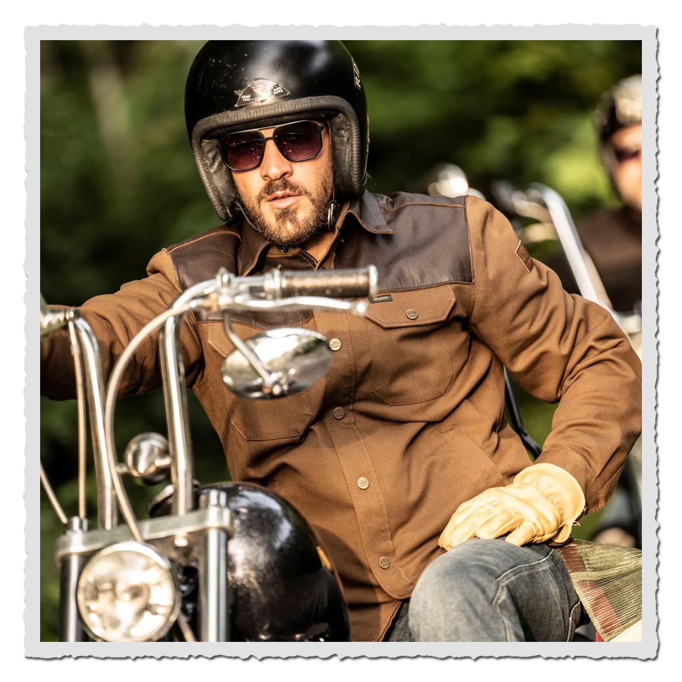 Motorradfahrer mit Helm und Sonnenbrille auf Bike, trägt braunes Rider Shirt, grüner Hintergrund im Abendlicht.